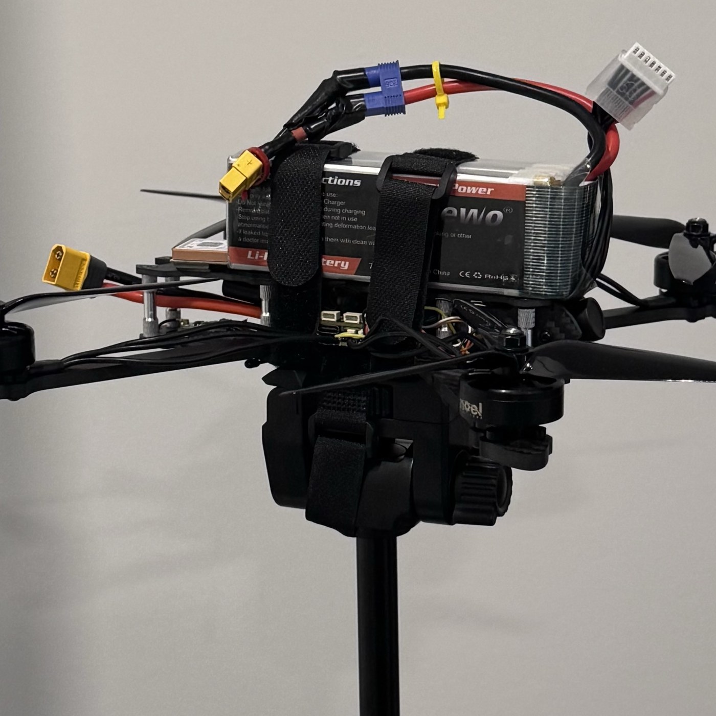 A seven-inch FPV quadcopter on a studio stand: a carbon frame with fixed propellers, a strapped lithium battery on top, and exposed wiring and connectors.