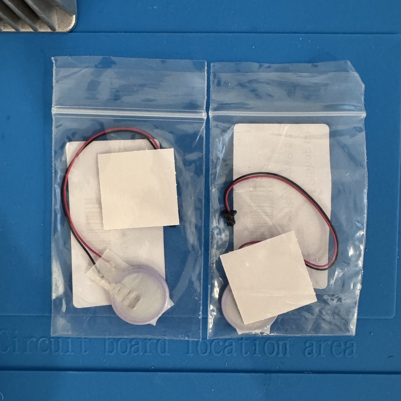 Two small rechargeable lithium coin cells with red and black leads, one with a two-pin connector, each in a clear bag on a blue work mat.